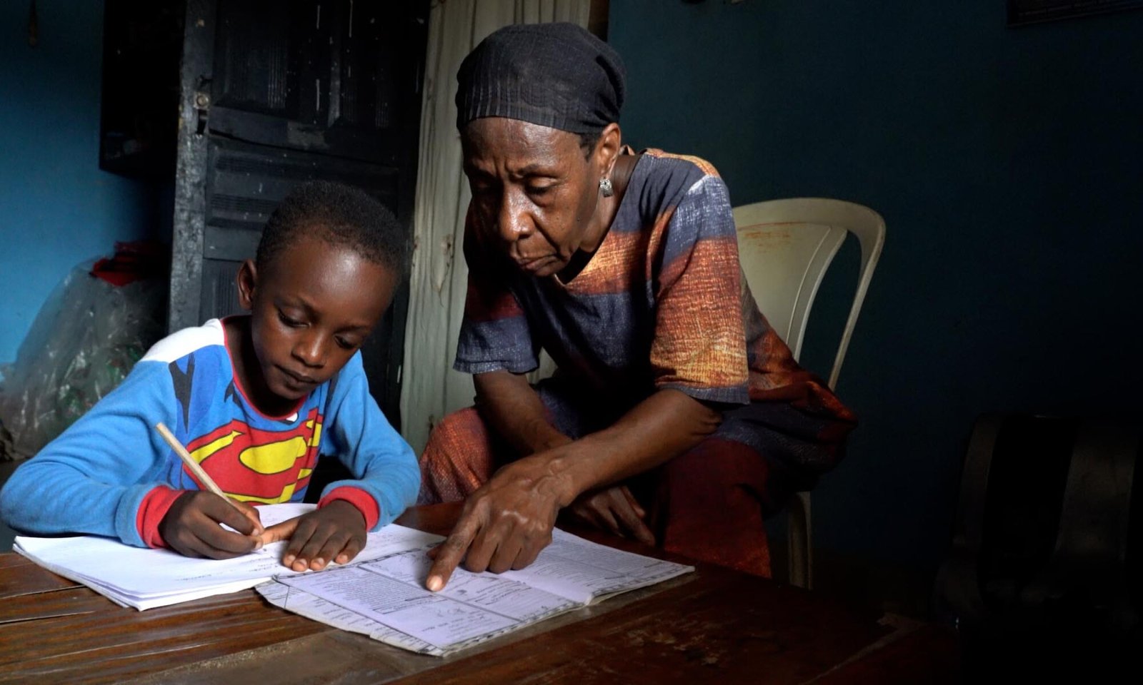 A young boy draws with a pencil at a wooden table while an elderly woman watches over him in a dim blue-walled room, guiding his work.
