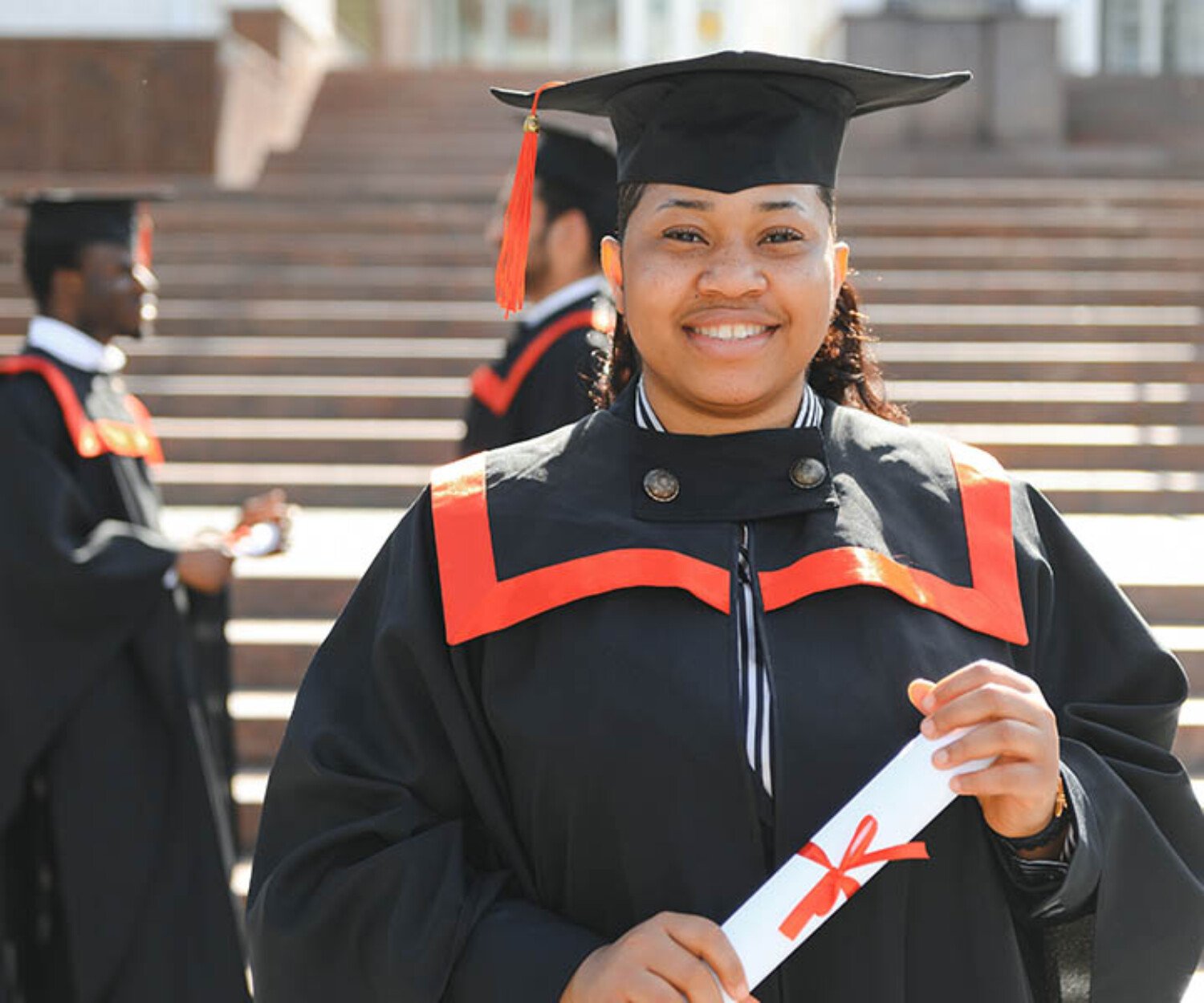 Graduate in cap and gown holding a diploma on campus steps, smiling at the camera