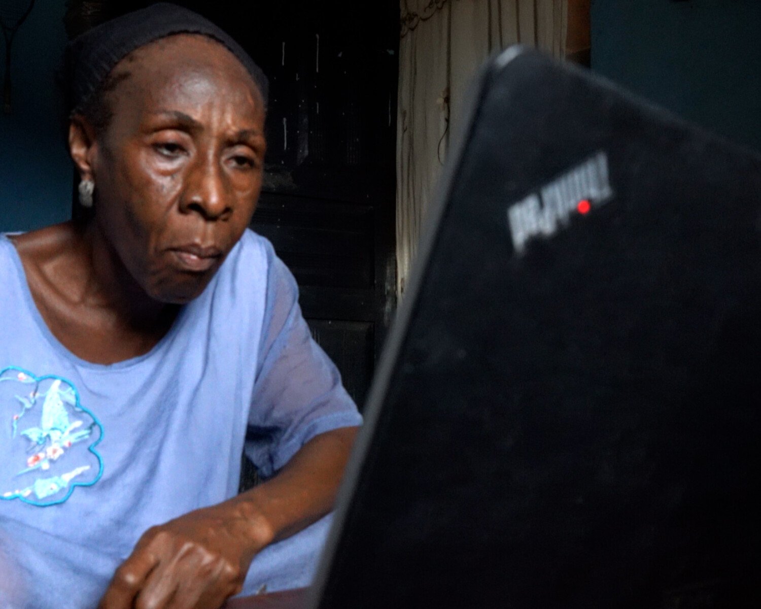 Older woman in a light blue shirt works at a laptop in a dim room, focused on the screen.