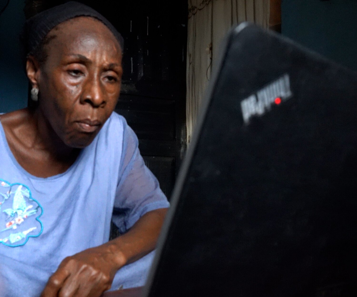 Older woman in a light blue shirt works at a laptop in a dim room, focused on the screen.