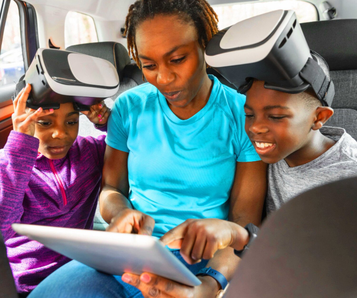Family in a car wearing VR headsets, looking at a tablet together in the back seat.