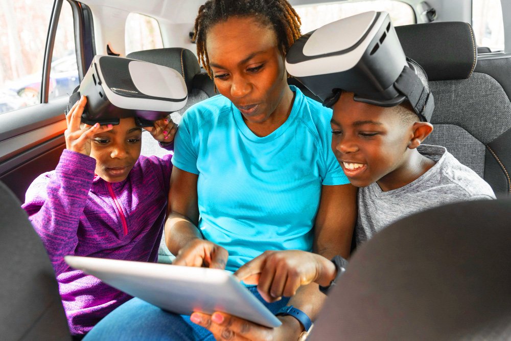 Family in a car wearing VR headsets, looking at a tablet together in the back seat.