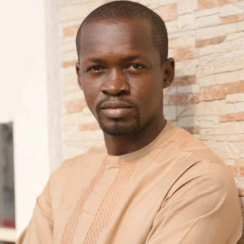 Portrait of a man in a beige traditional outfit leaning against a brick wall, looking at the camera.
