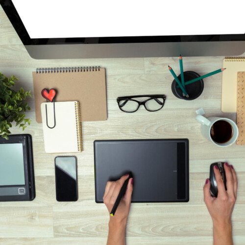 Top-down view of a workspace: a person draws on a graphics tablet with a stylus, surrounded by a monitor, smartphone, glasses, coffee, plant, and notebooks.