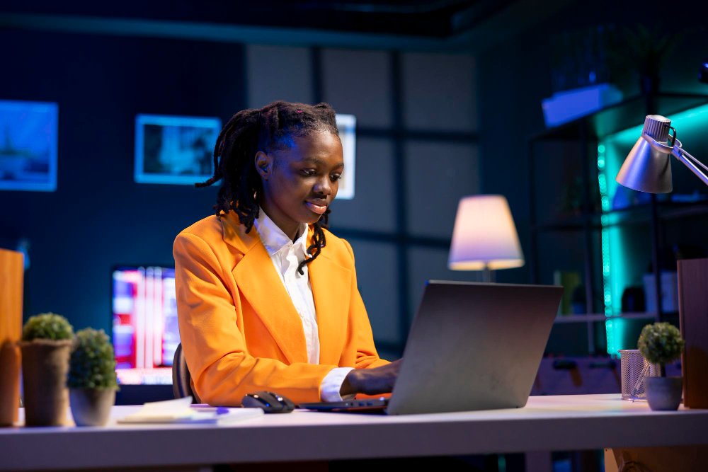 Professional woman in an orange blazer typing on a laptop at a desk in a modern office setting with plants nearby.